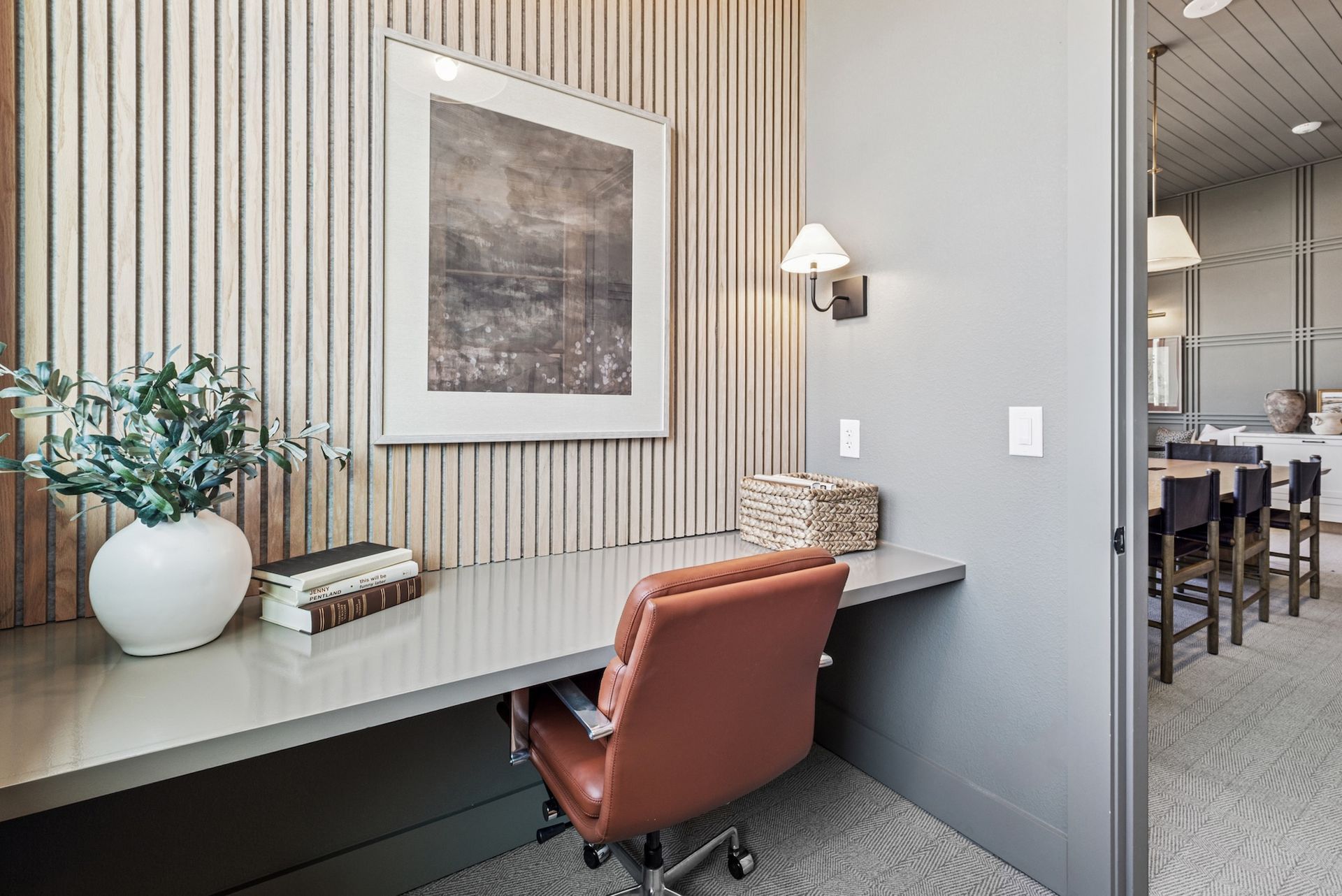 Home office with desk, chair, artwork, and white vase with greenery. Adjacent to dining area.