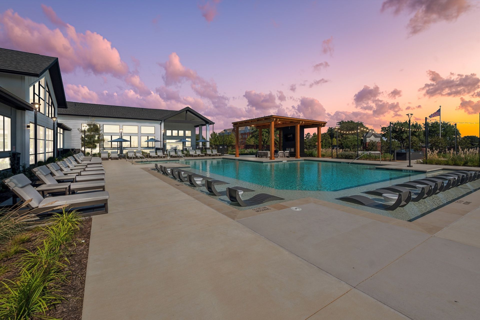 Pool with lounge chairs at dusk; modern building, pergola, and colorful sky.