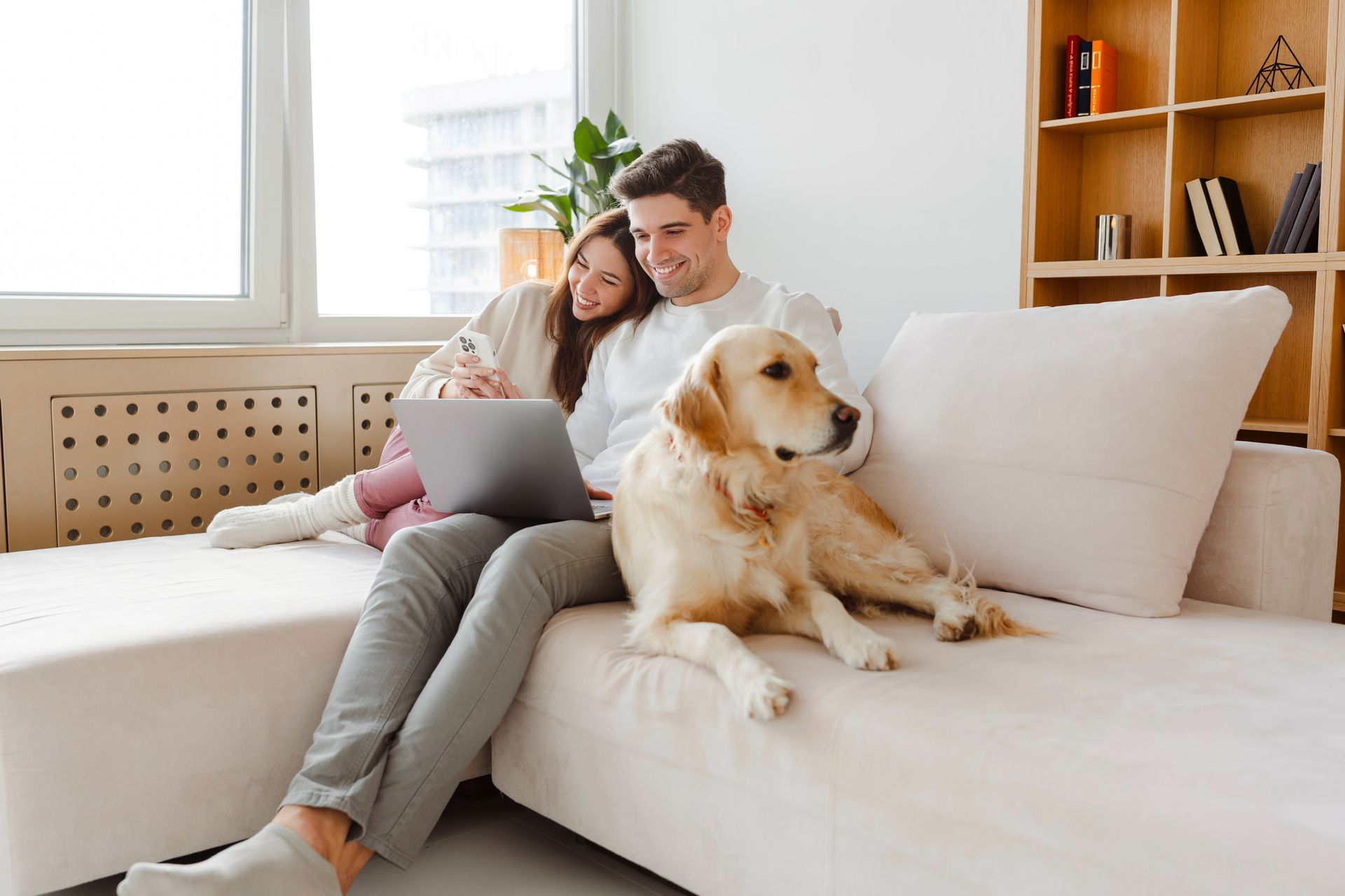 A man and woman are sitting on a couch with a dog and a laptop.