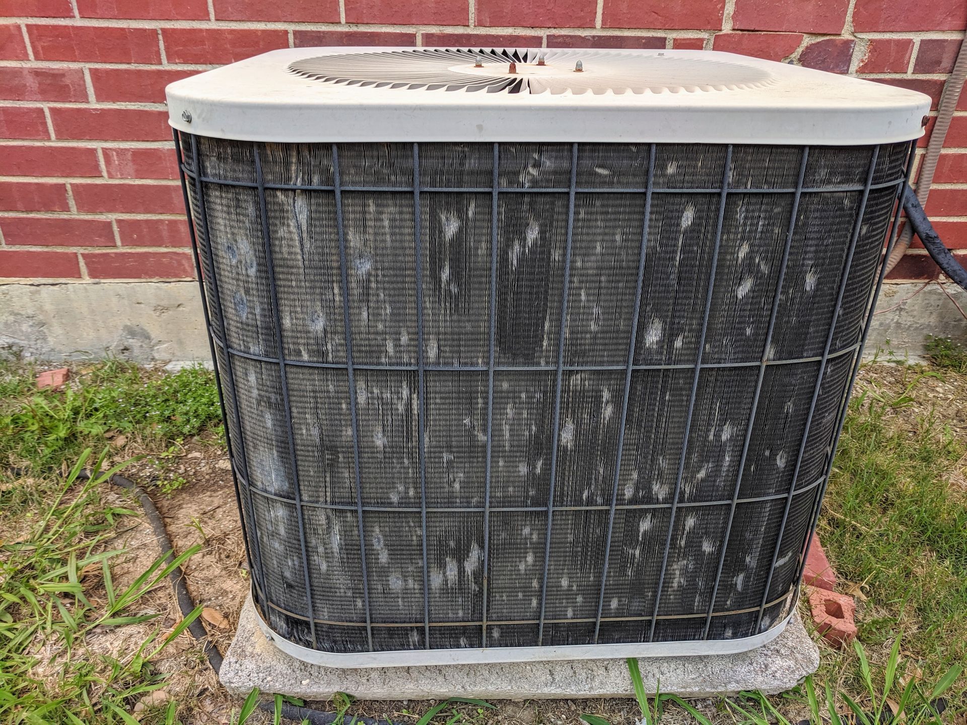 Air conditioner unit with clogged, dusty fins sits on a concrete pad near a brick wall and grass.