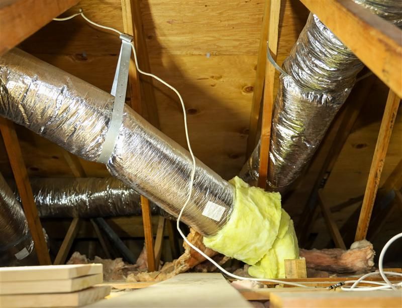HVAC ductwork in an attic. Insulated silver ducting, yellow insulation at the end, supported by metal straps.
