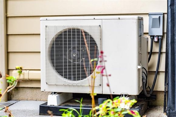 White AC unit on a black base beside a house, with electrical box, and blurry flowers.