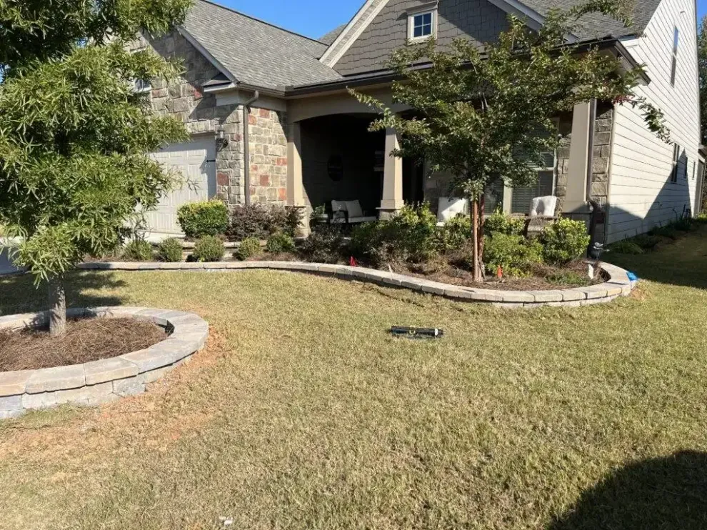 A tan brick house with a front porch, landscaping with stone borders, and trees in the foreground under a clear blue sky.