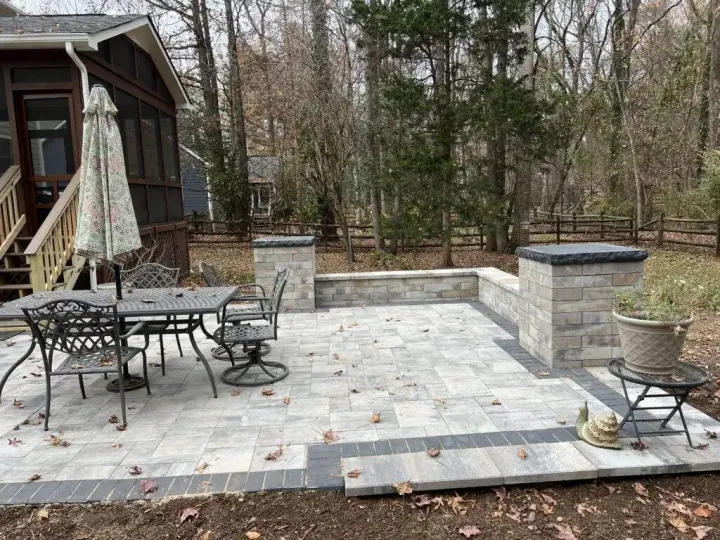 A backyard patio with light stone pavers, a dining set, and low retaining walls, situated beside a house with a deck.