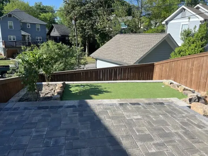 A backyard featuring a stone paver patio leading to a rectangular artificial turf area bordered by rocks and wooden fences.