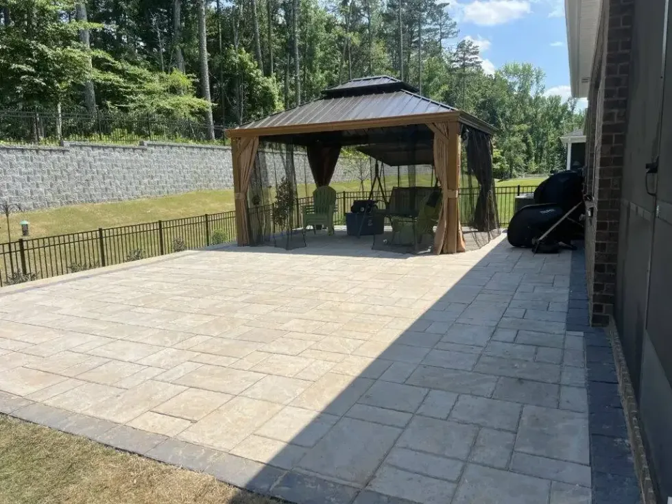 A backyard patio featuring a large stone-paver surface, a wooden gazebo with furniture, and a stone retaining wall.
