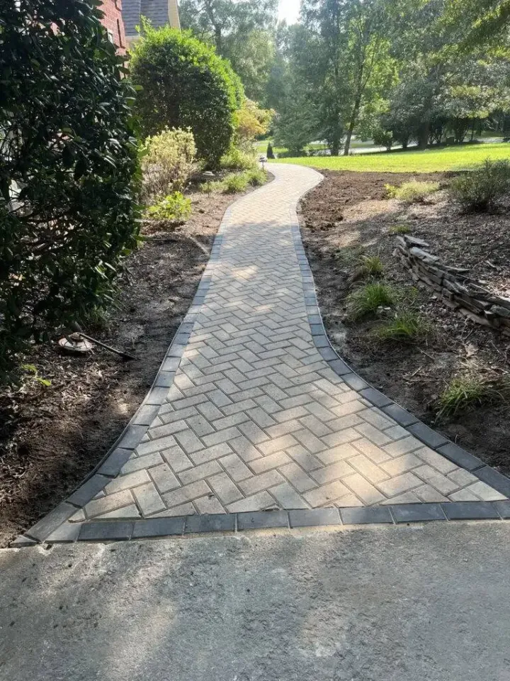 A herringbone-patterned stone walkway winds through a garden area with green trees and a grassy lawn in the background.