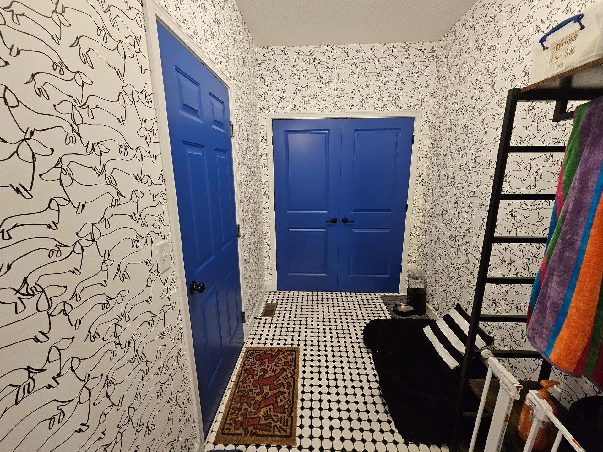 Narrow hallway with patterned wallpaper, blue doors, black and white floor tiles, and a ladder with towels.