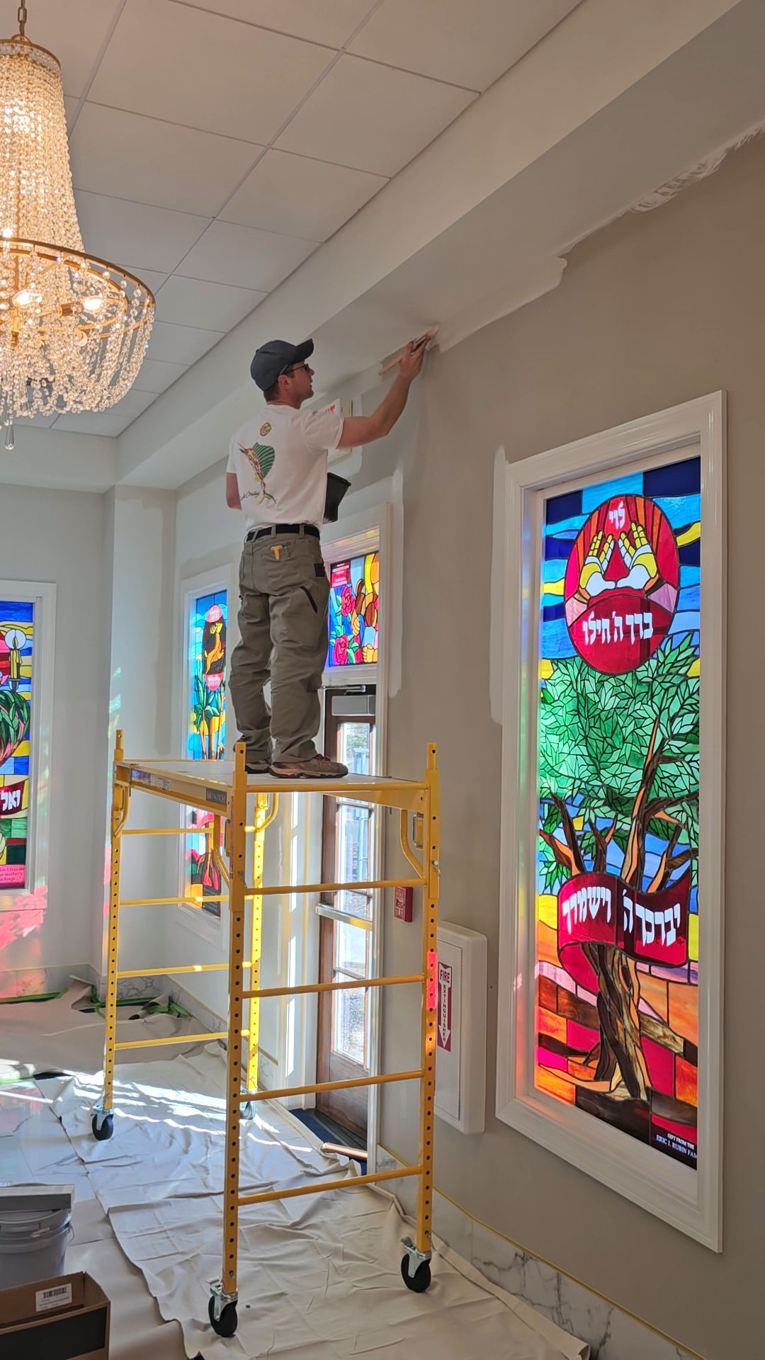 Painter on a scaffold brushes white paint onto crown molding in a room with stained-glass windows and a chandelier.