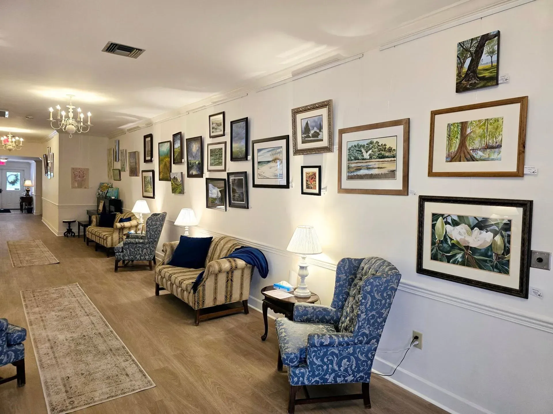 Long hallway with framed art, blue patterned chairs, side tables, and a lit lamp in a cozy waiting area.