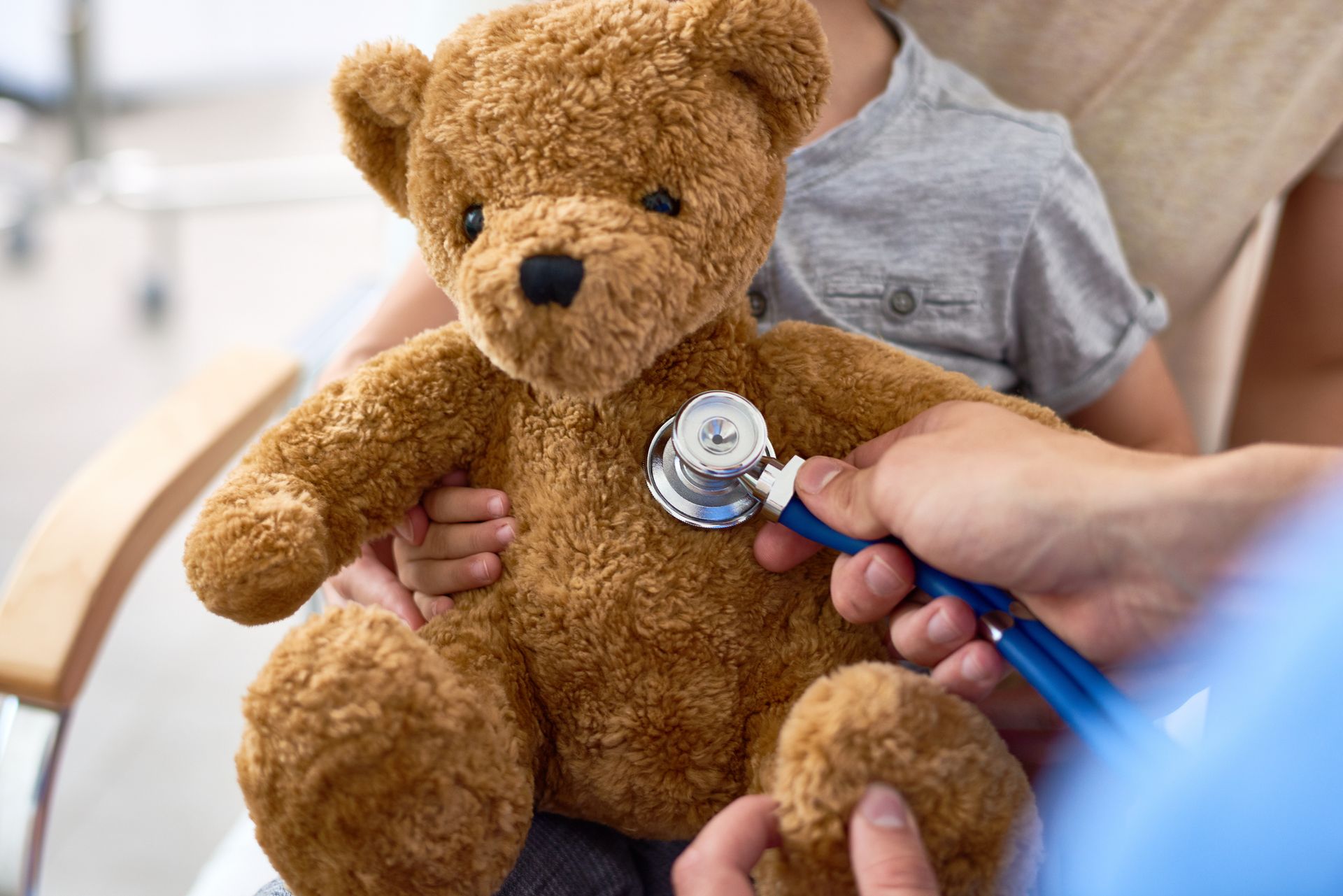 Doctor using a stethoscope to examine a teddy bear with a child present in a medical setting.