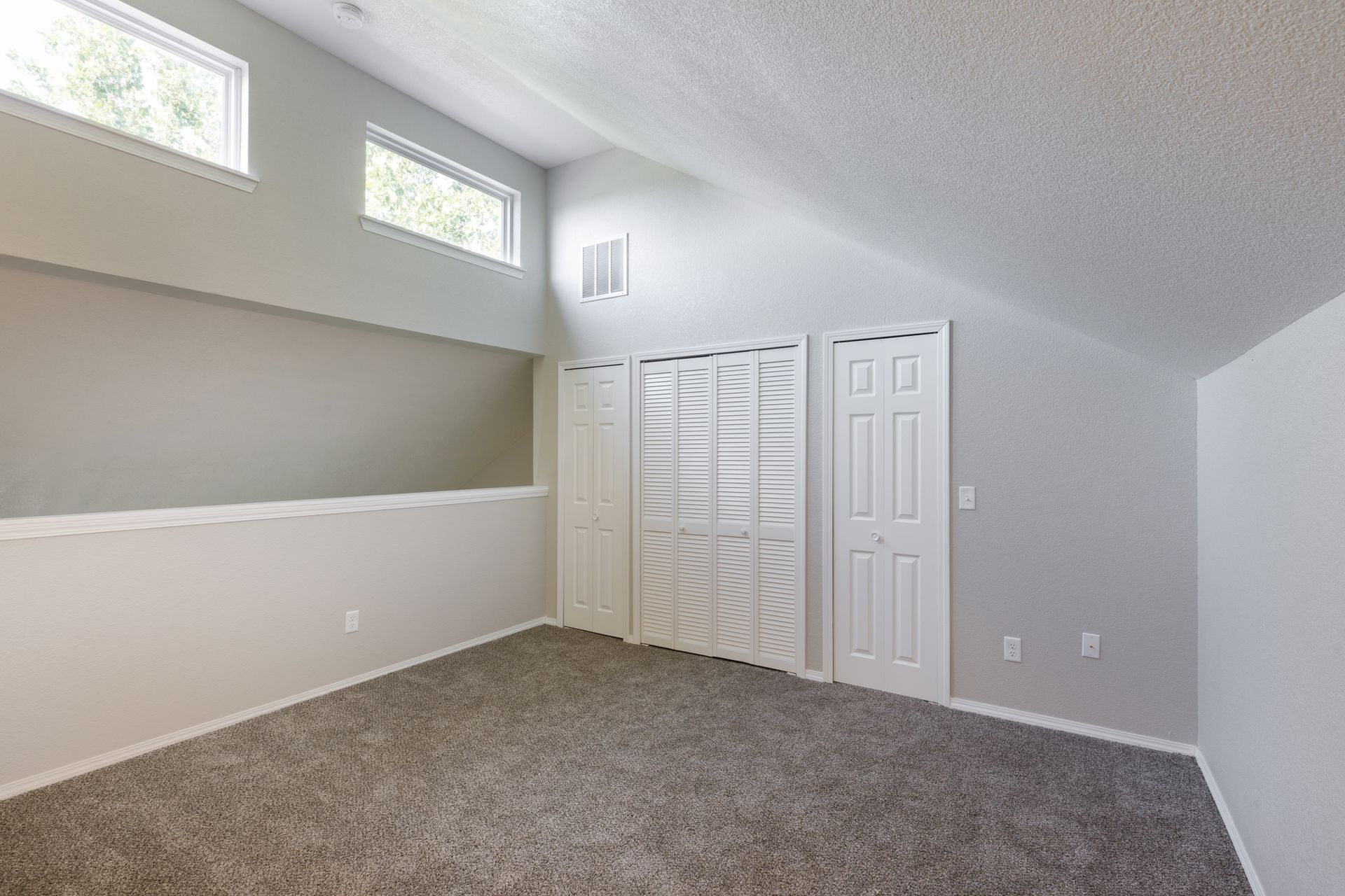 Empty bedroom with sloped ceiling, carpet, white doors, and small windows.
