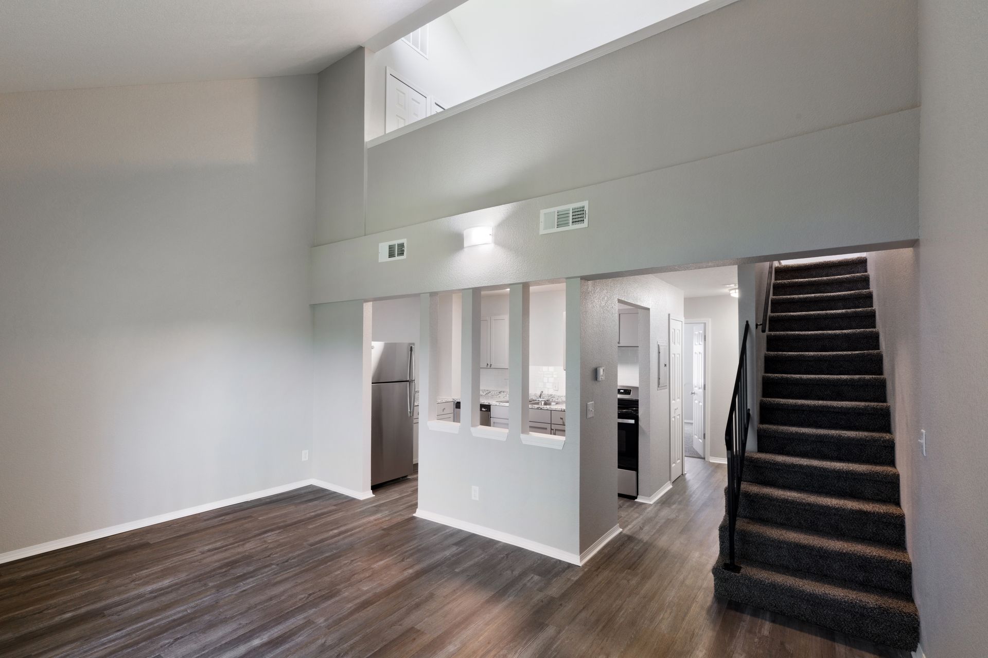 Living room with gray walls, kitchen area, and staircase. Dark hardwood floors and tall ceilings.