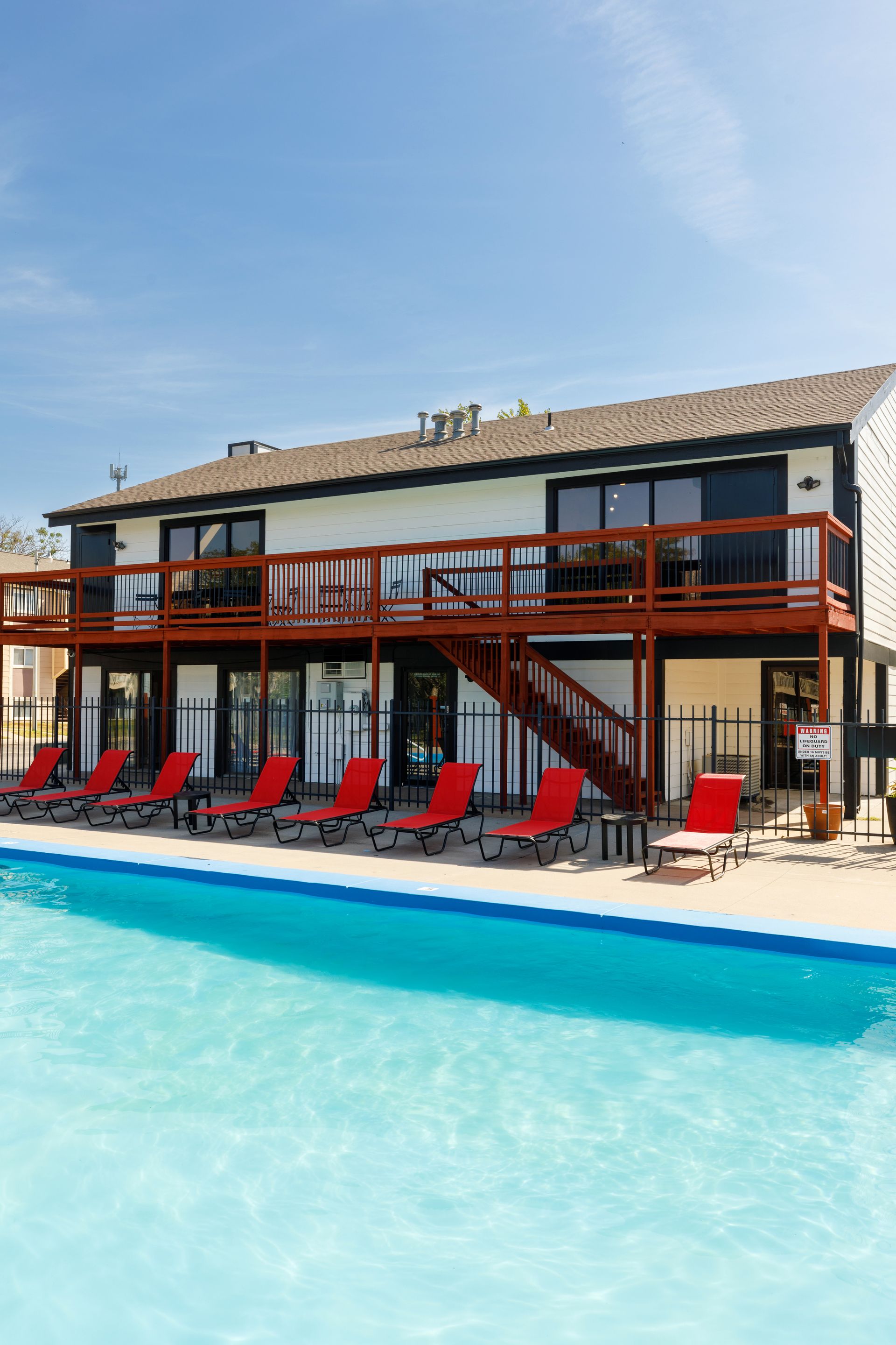 Two-story building with a pool, red lounge chairs, and a clear blue sky.