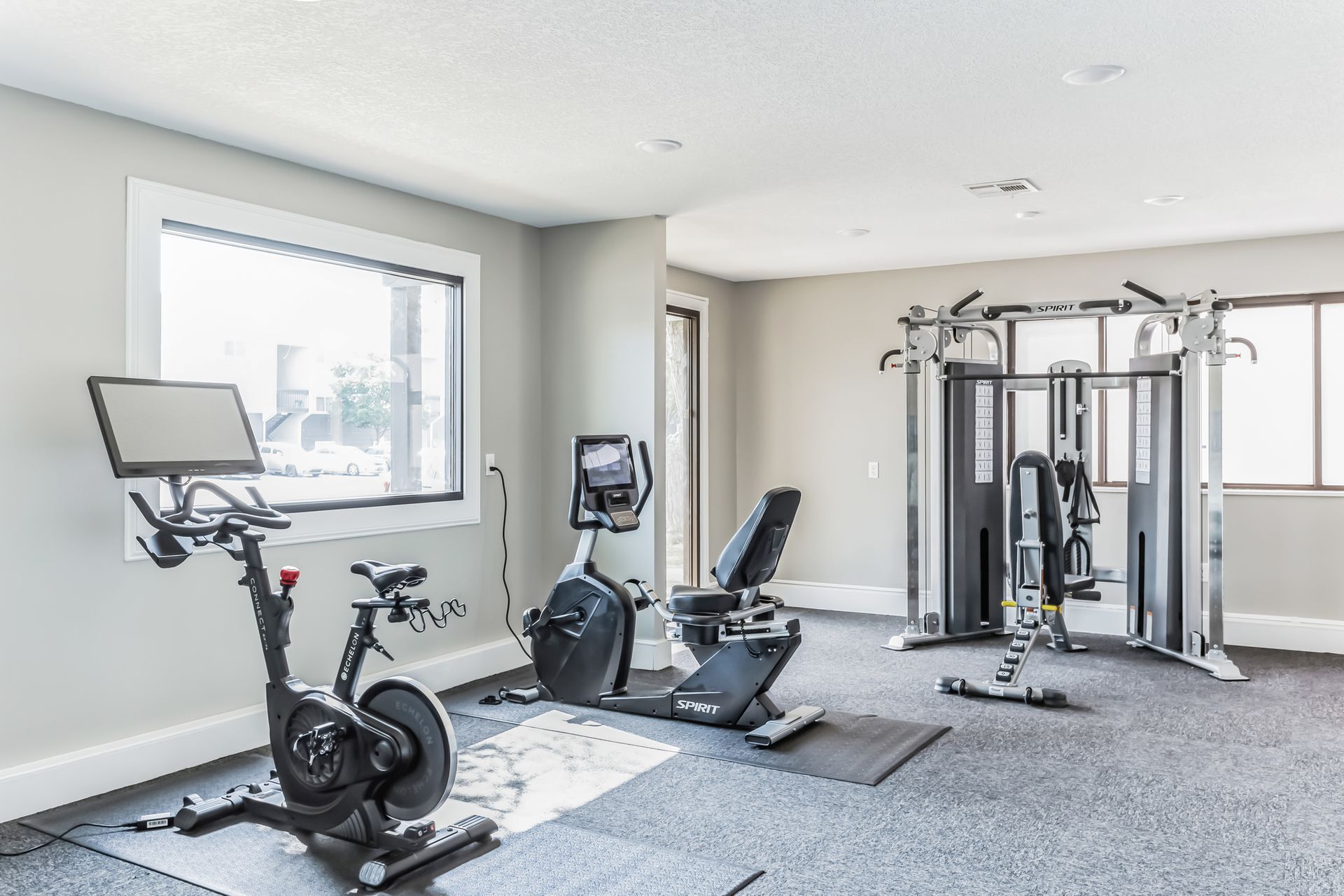 Gym interior with exercise bikes, weight machine, and recumbent bike. Grey walls, gray floor.