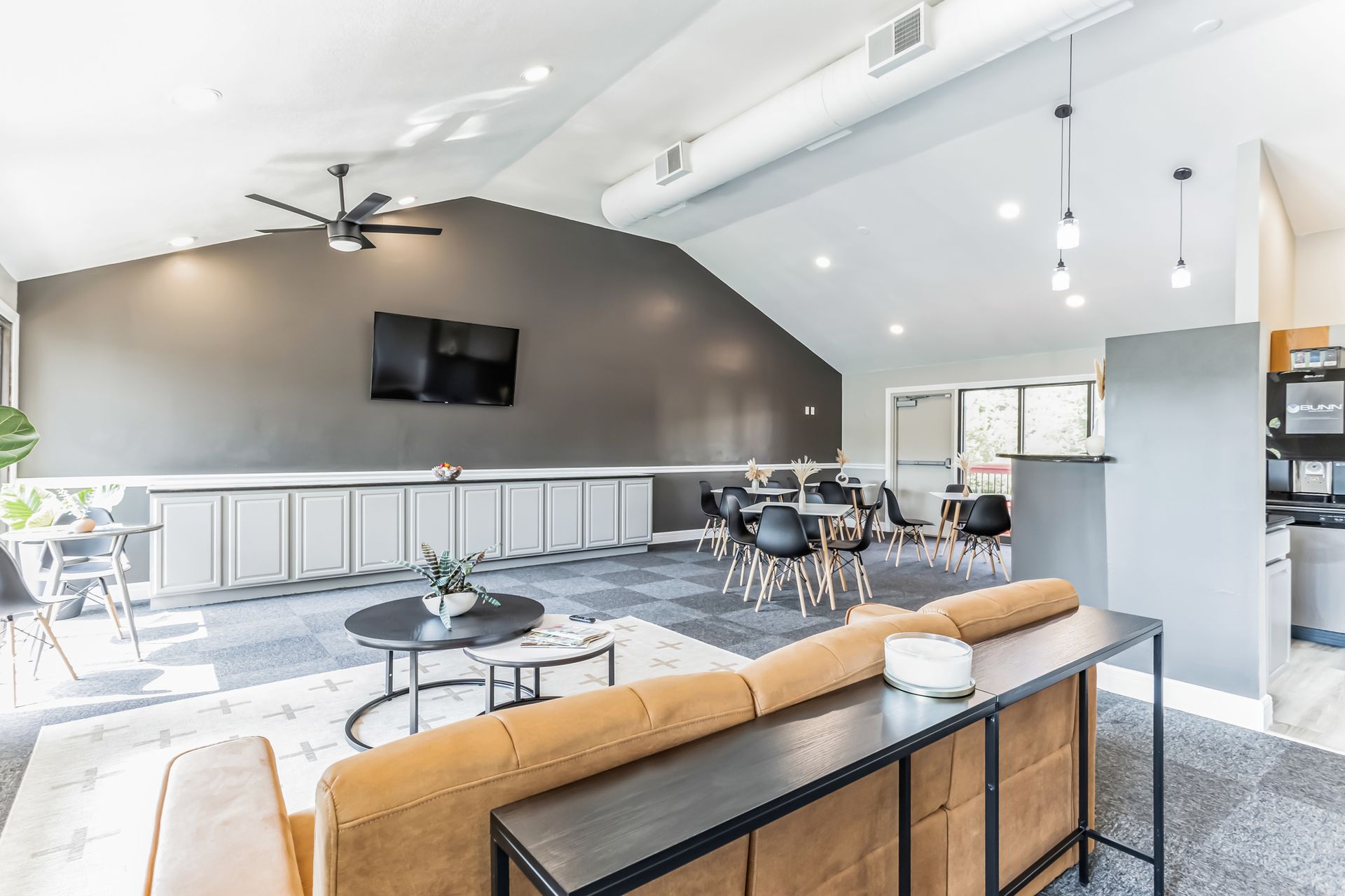 Modern lounge area with tan couch, black tables, and chairs; grey walls and ceiling; a TV.