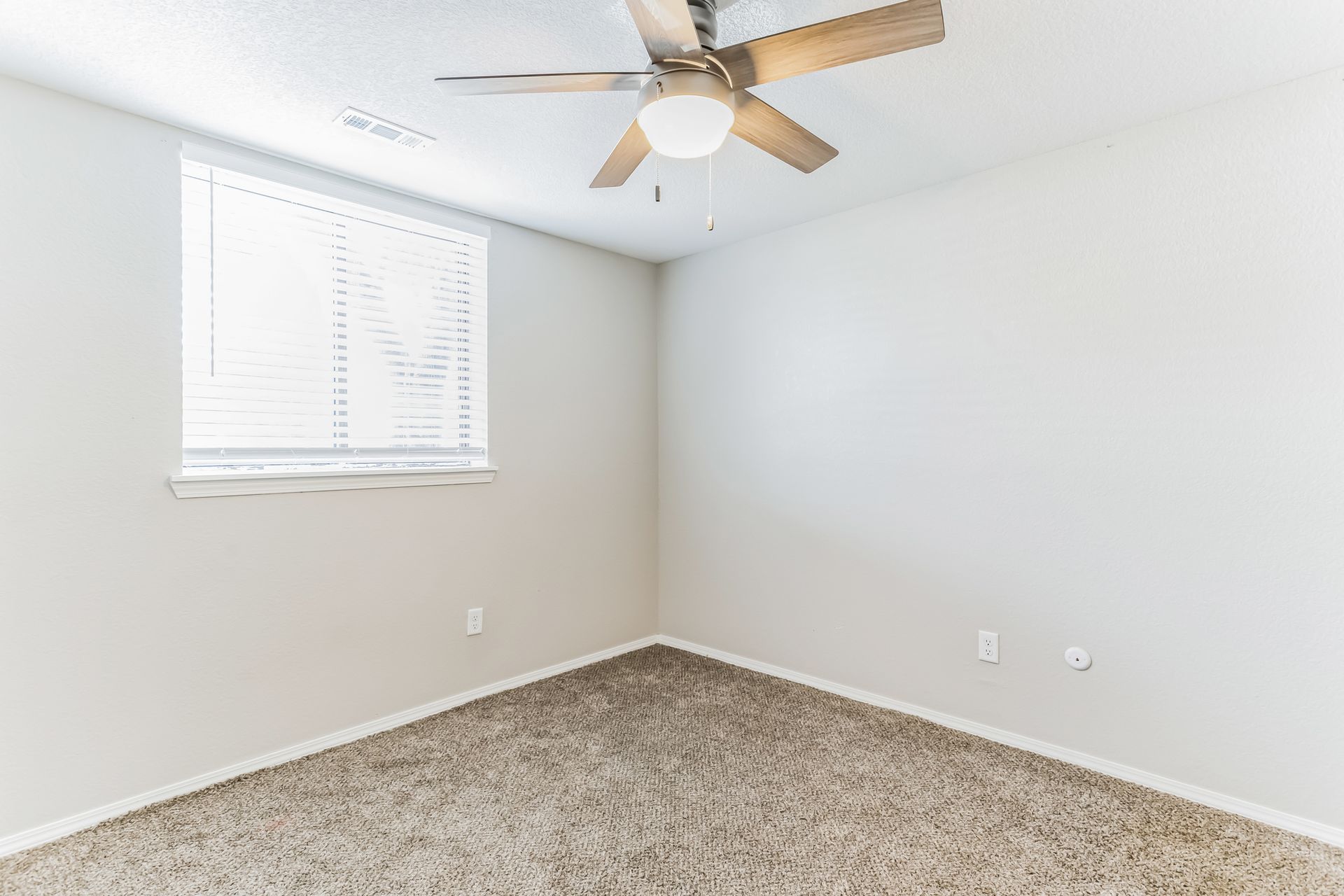 Empty beige bedroom with carpet, window with blinds, and ceiling fan.