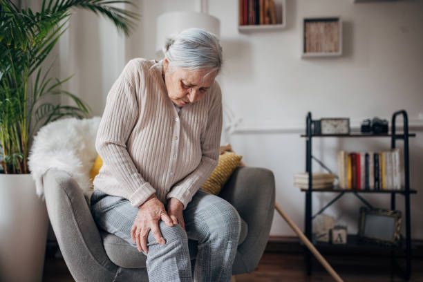 An Elderly Woman Is Sitting In A Chair Holding Her Knee In Pain — Chicago, IL — Chicagoland Home Physicians