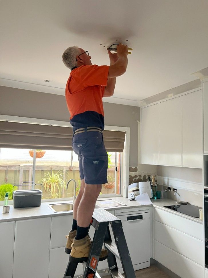 A Man in an Orange Shirt is Standing On A Ladder Replacing A Smoke Alarm — Rob Fritsch Electrical in Wagga Wagga, NSW