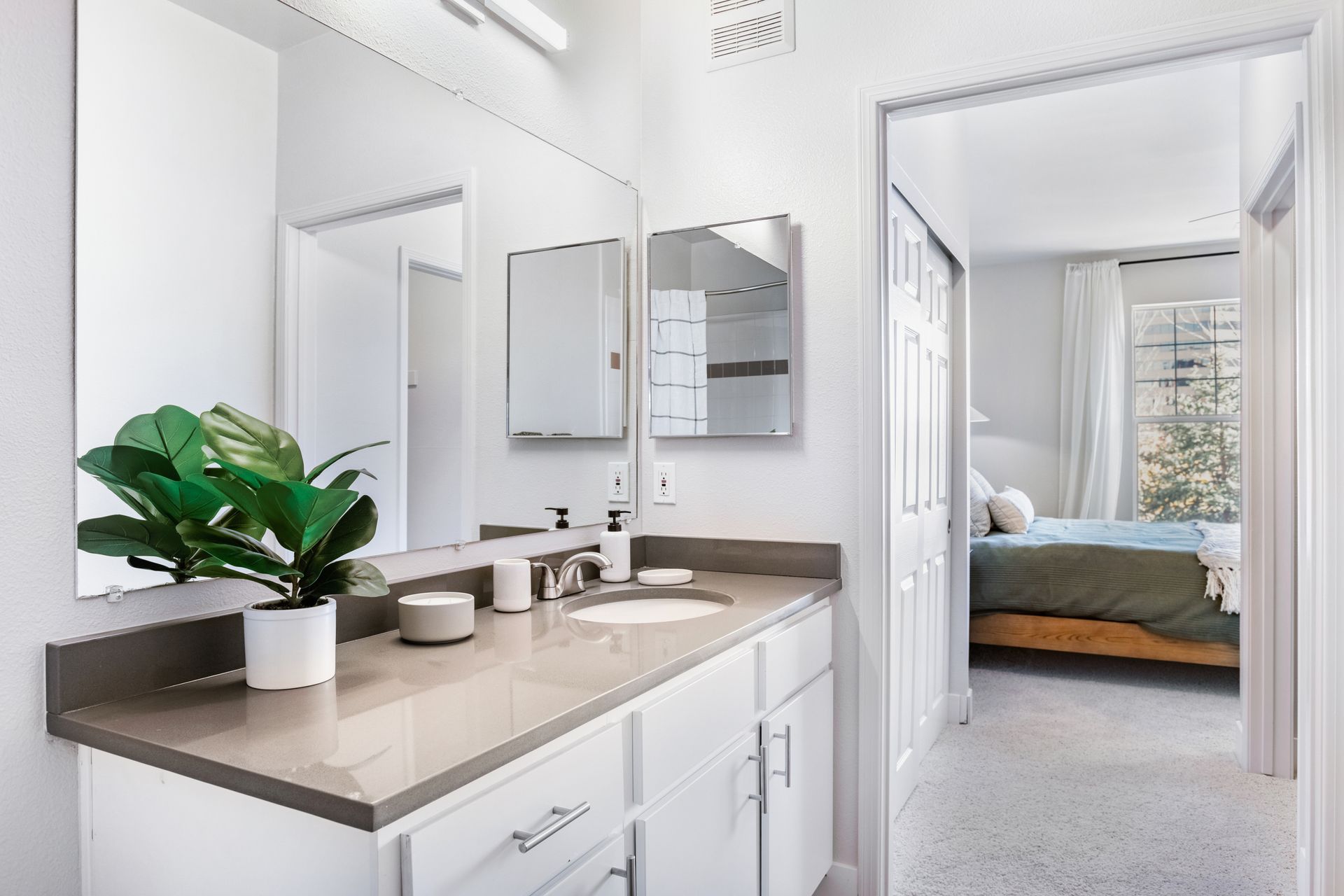 Bathroom with a white vanity, large mirror, and a view into a bedroom with a bed and window.