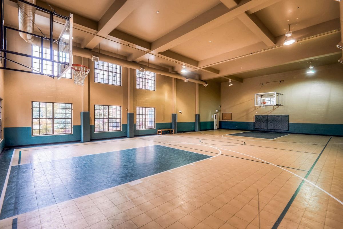 Empty school gymnasium with basketball hoops, blue and tan floor, windows, and beige walls.