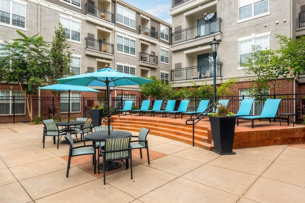 Outdoor apartment patio with lounge chairs and tables. Teal umbrella and chairs on a brick deck.