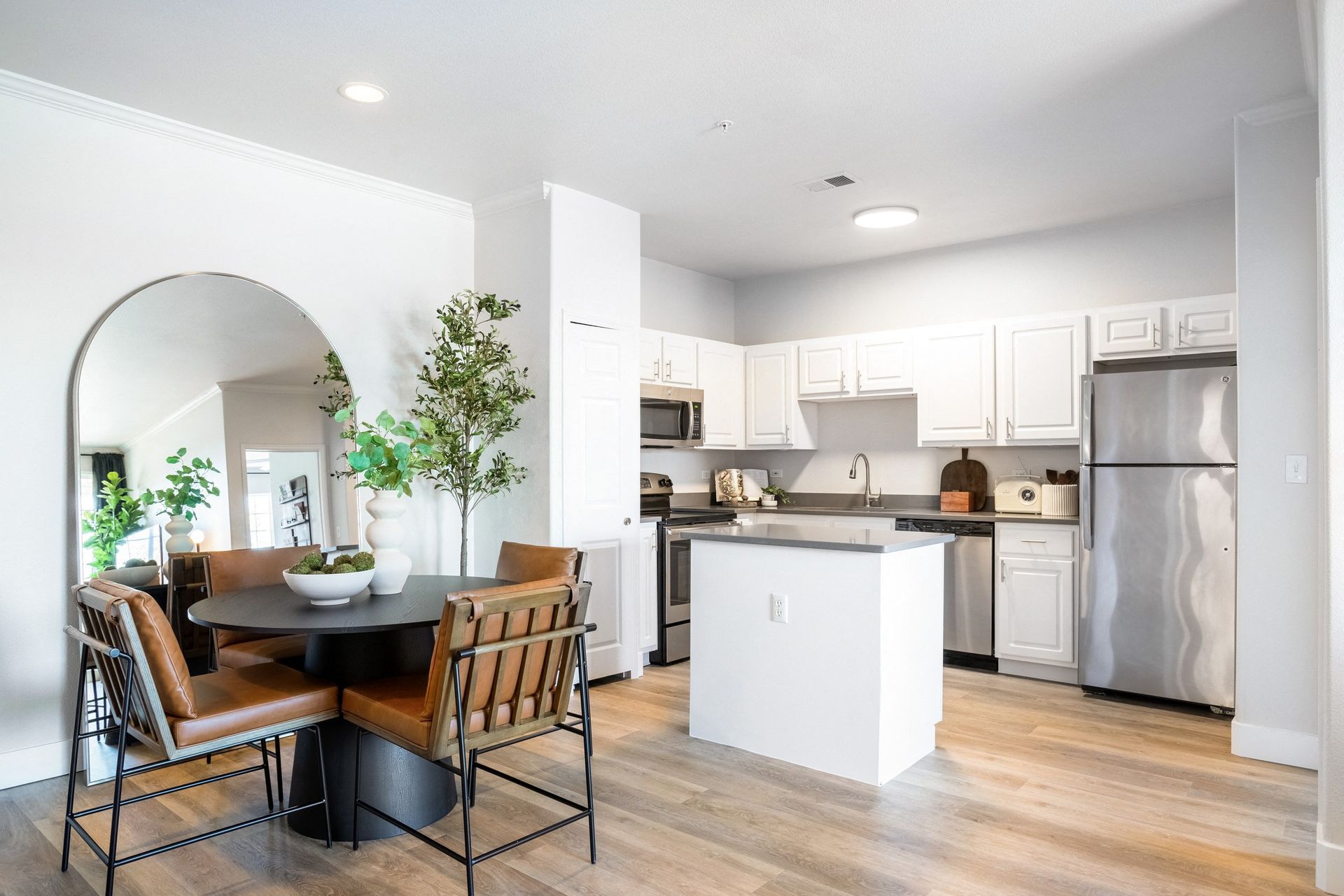 Open-concept kitchen/dining area: round table with leather chairs, arched mirror, white cabinets, stainless steel appliances, wood floors.