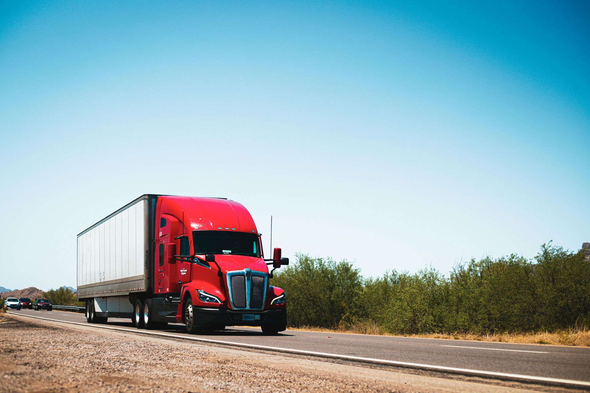 Red semi-truck driving on a highway, under a clear blue sky.