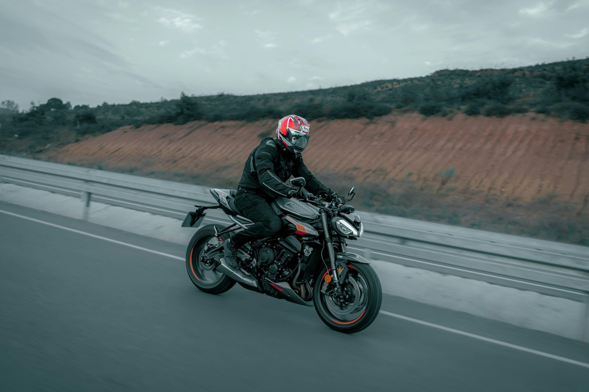 A motorcyclist in black gear and a red helmet rides a dark sportbike along a highway beside an orange-colored embankment.