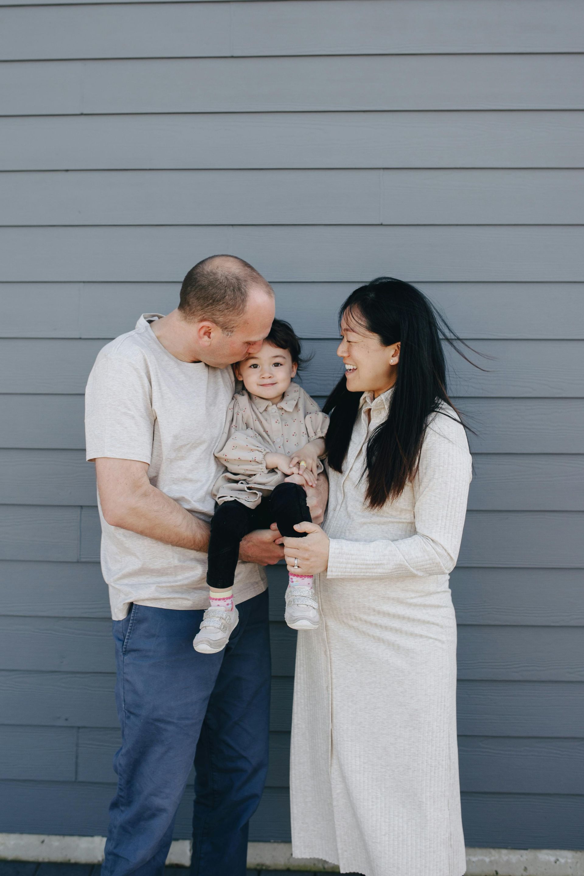 Family of three smiles in front of a blue wall; father kisses child's head, mother looks on.