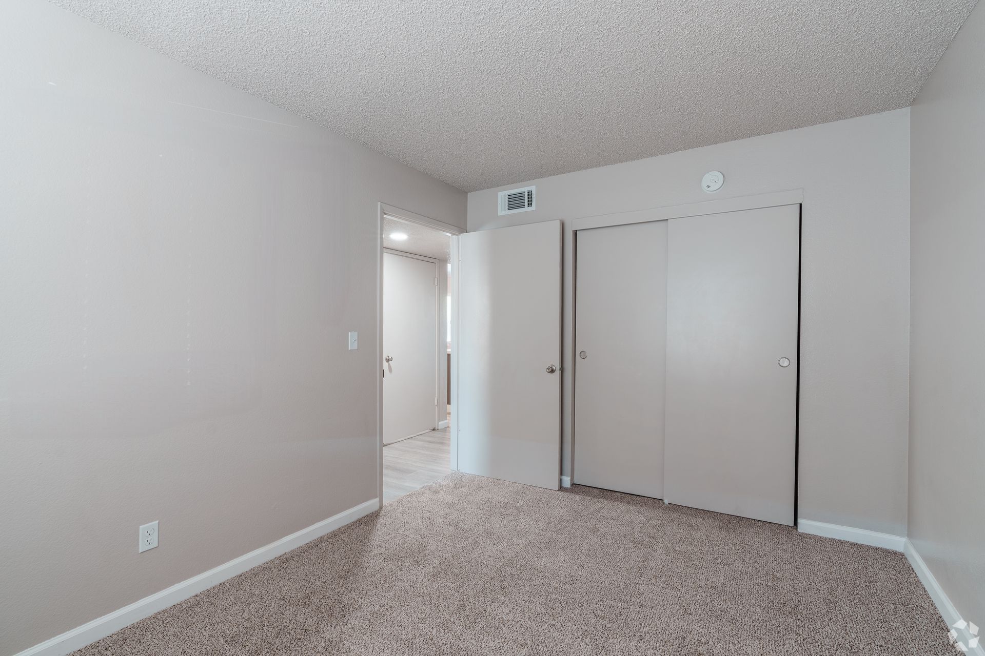 Empty bedroom with beige carpet, walls, and closet doors. A doorway to a hallway is on the left.