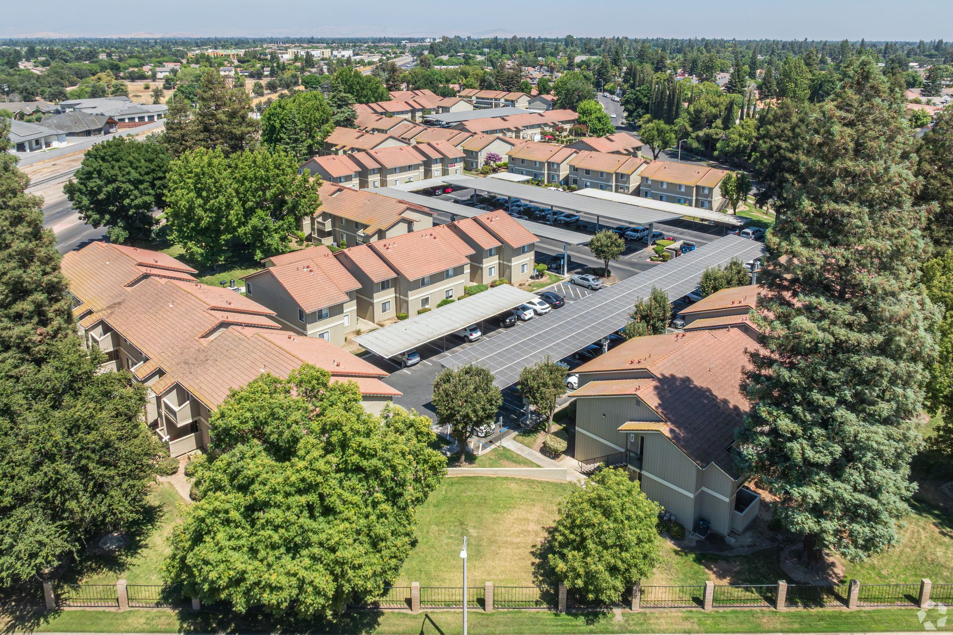 Aerial view of apartment complex with red-tiled roofs, surrounded by trees and covered parking.