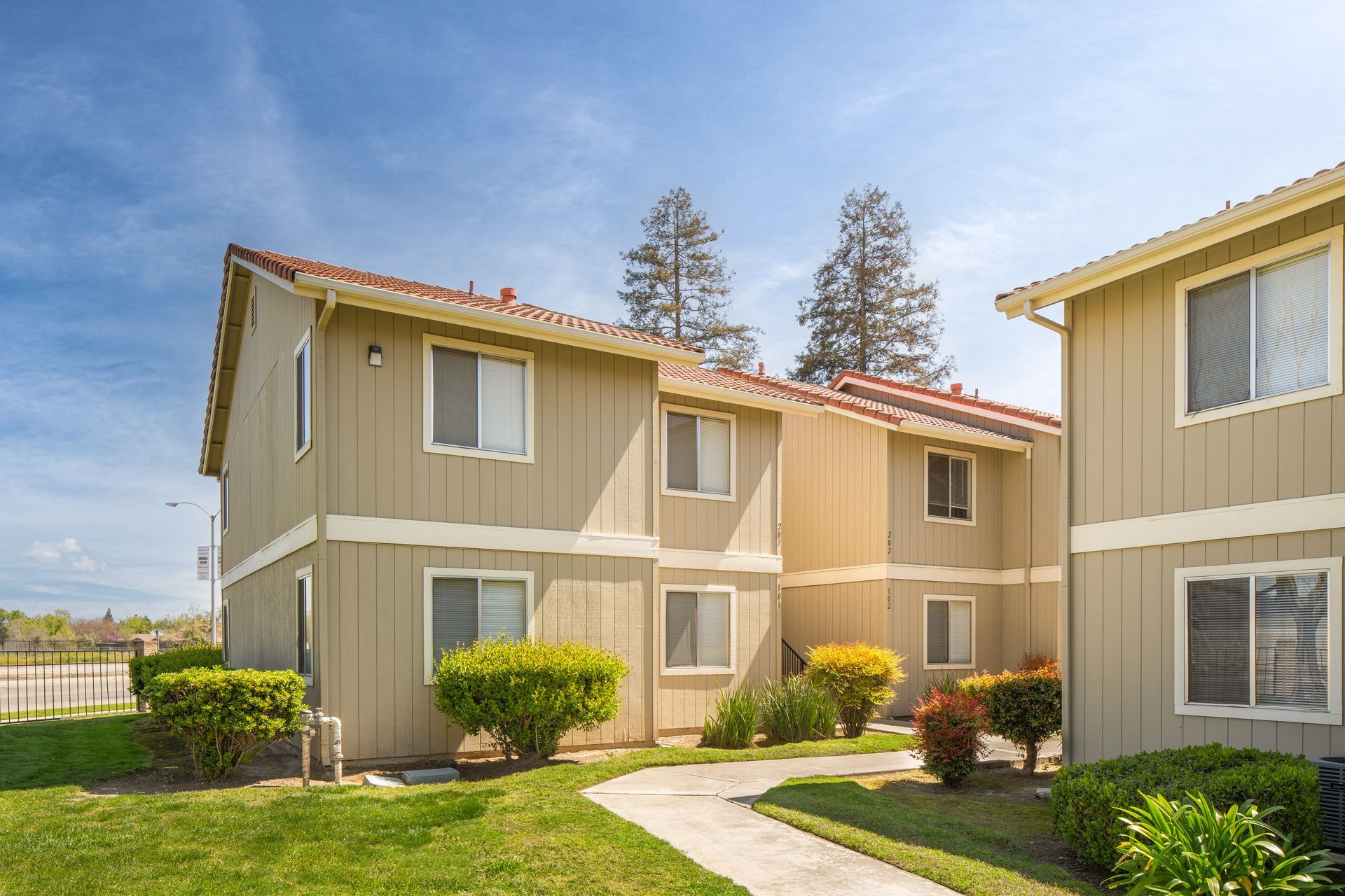 Tan two-story apartment buildings with tile roofs under a blue sky, walkway in front.