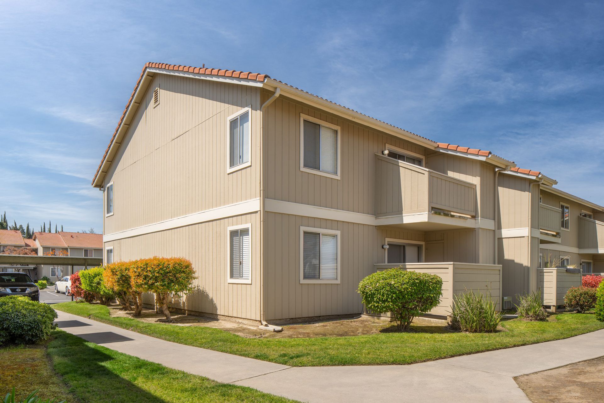 Two-story tan apartment building with red tile roof, green lawn, blue sky.