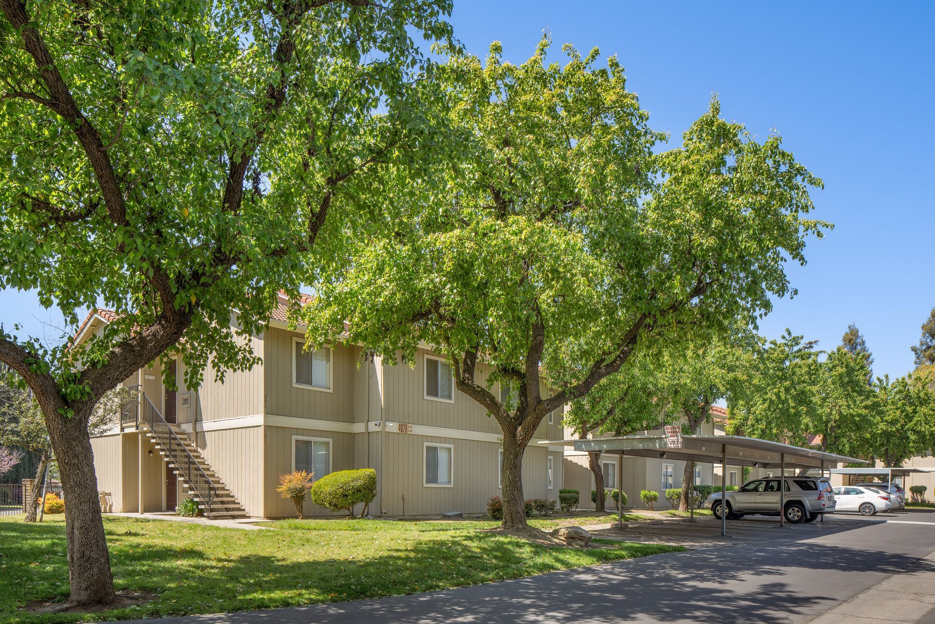 Two-story beige apartment building with trees, carport, and parked cars. Sunny day, clear blue sky.