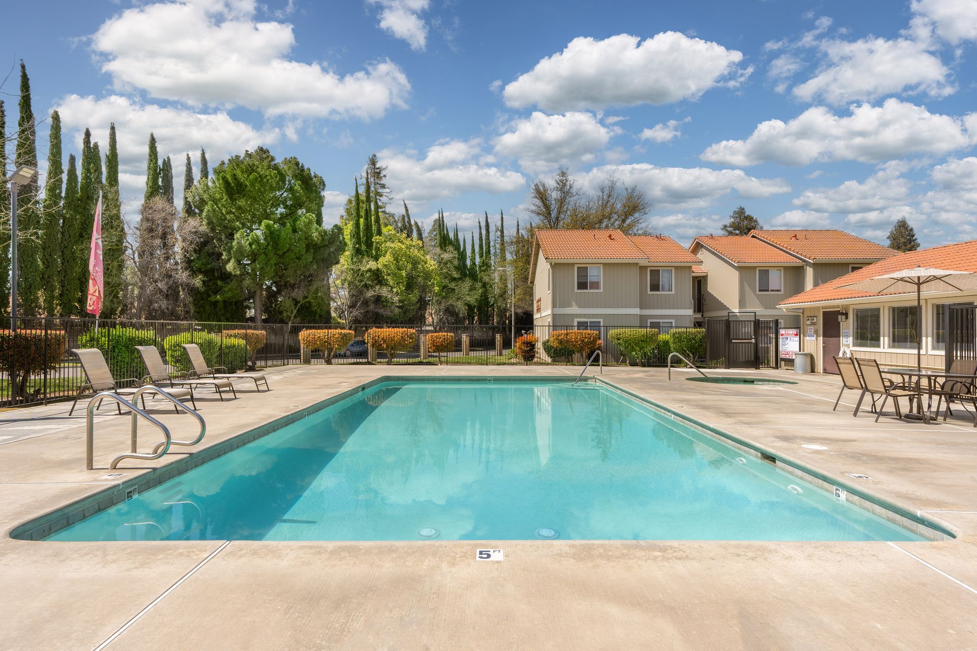 Swimming pool with lounge chairs, building, and trees under a blue sky.