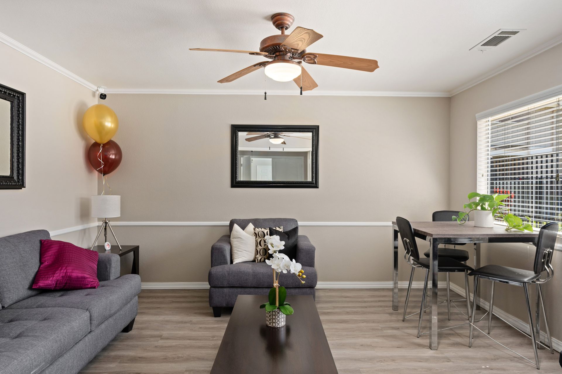 Living room with grey furniture, a mirror, and a dining table; brown ceiling fan; balloons.