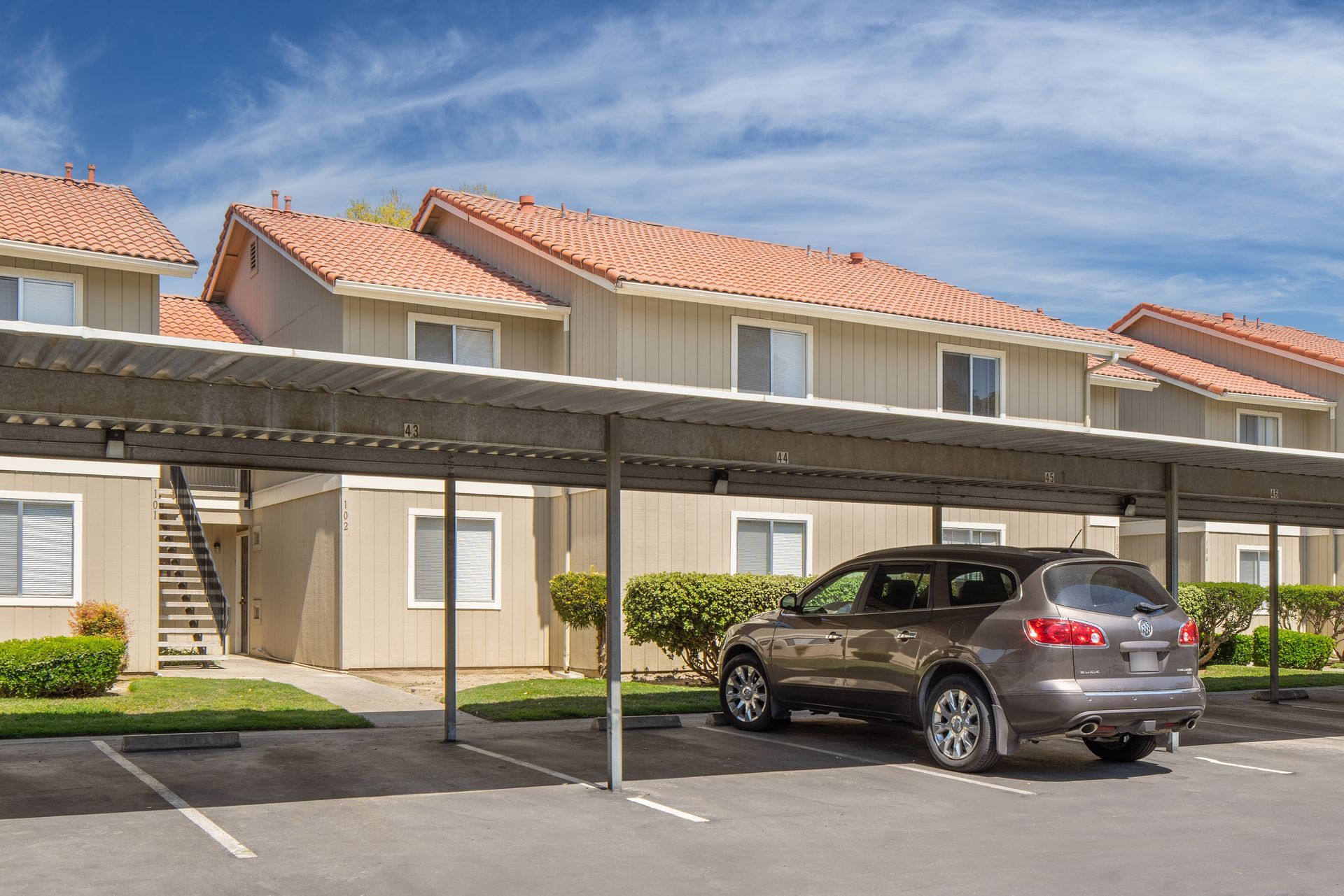 A brown SUV parked under a metal carport in front of a two-story apartment building with red-tiled roofs.