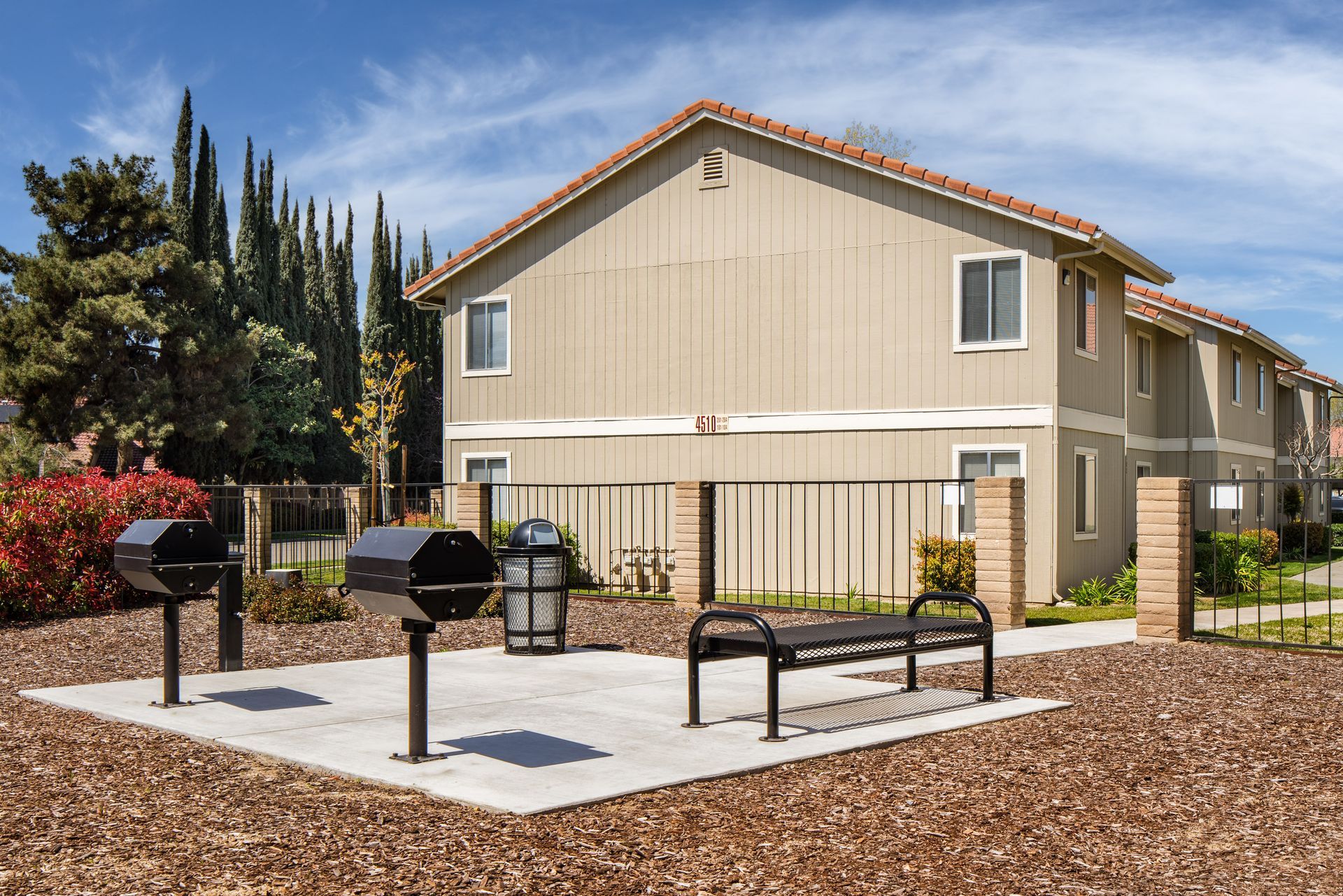 Two-story tan building with a patio featuring grills and a bench.