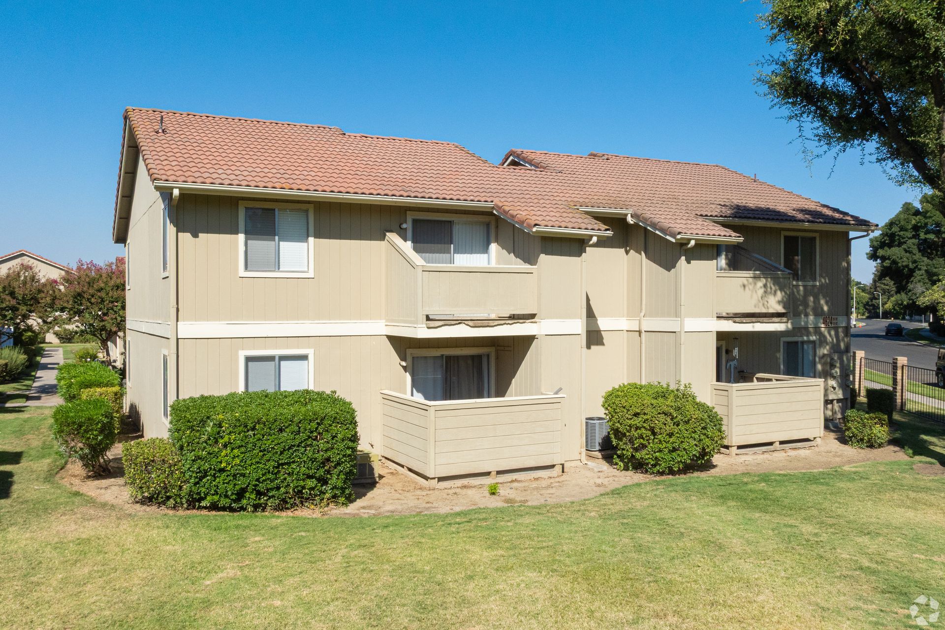 Two-story beige apartment building with red-tiled roof, green grass, and shrubs.