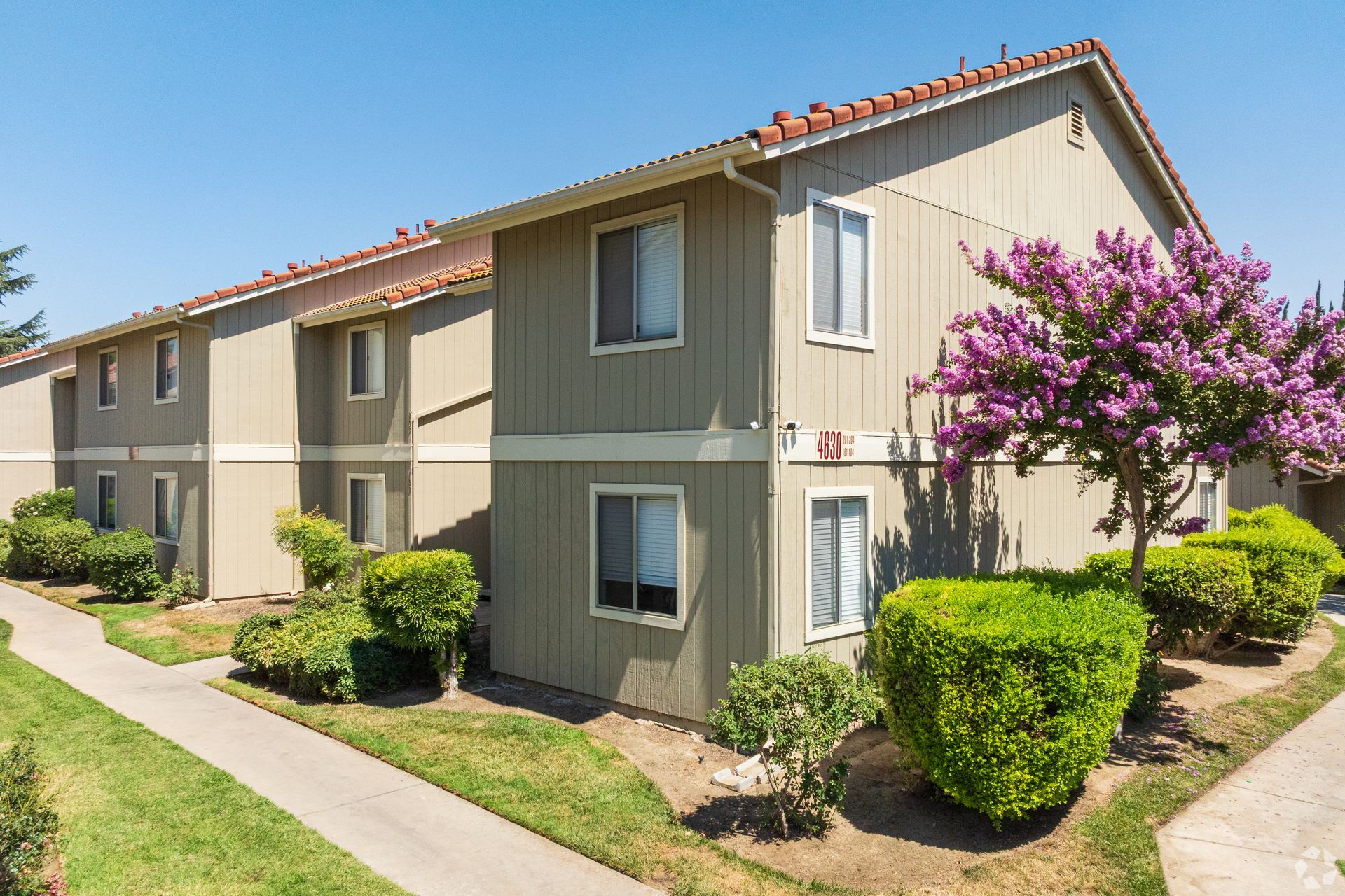 Apartment building with light beige siding, red tile roof, and landscaping under a clear sky.