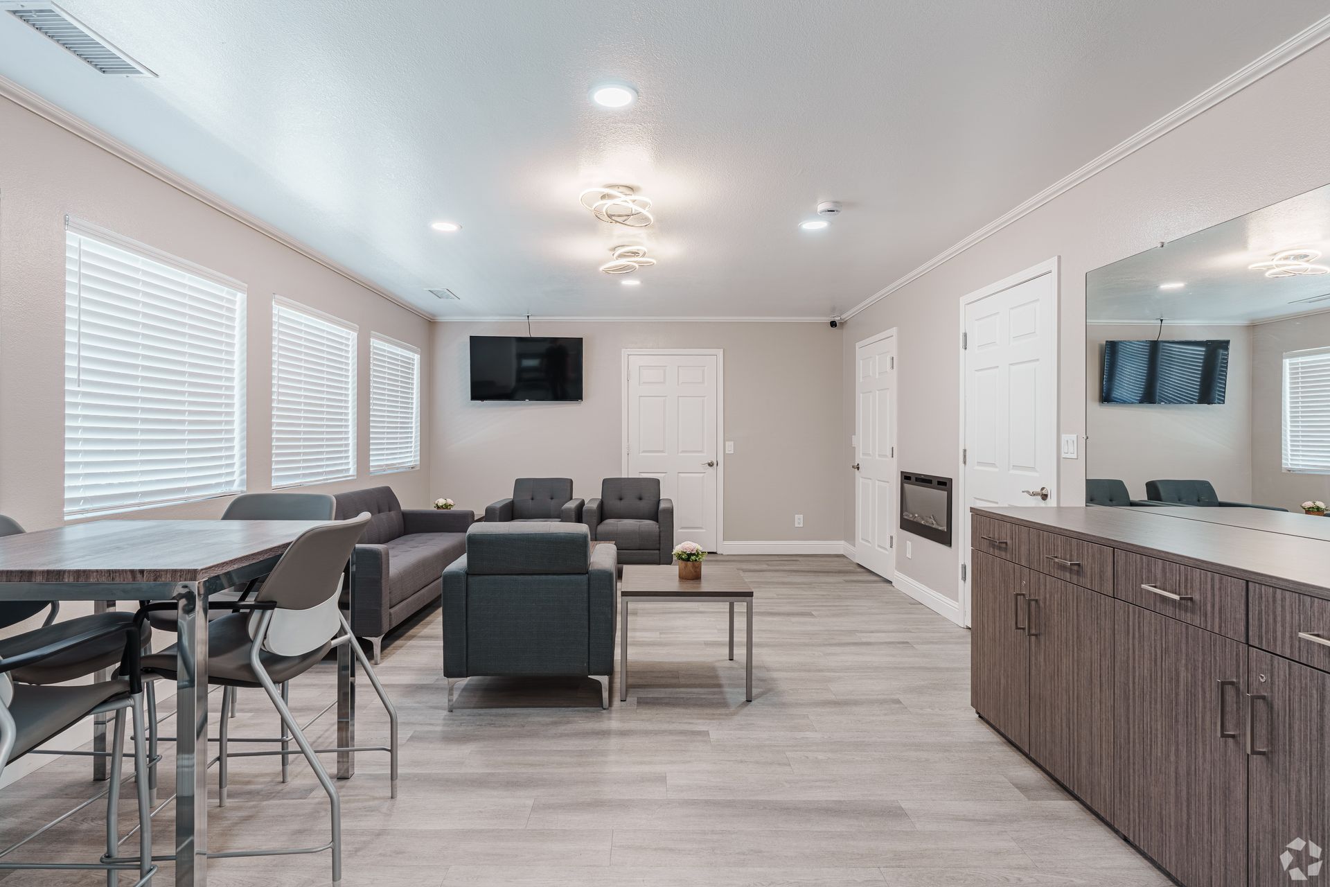 Spacious lounge with gray furniture, dining table, and large windows. Includes a kitchen counter and two TVs.