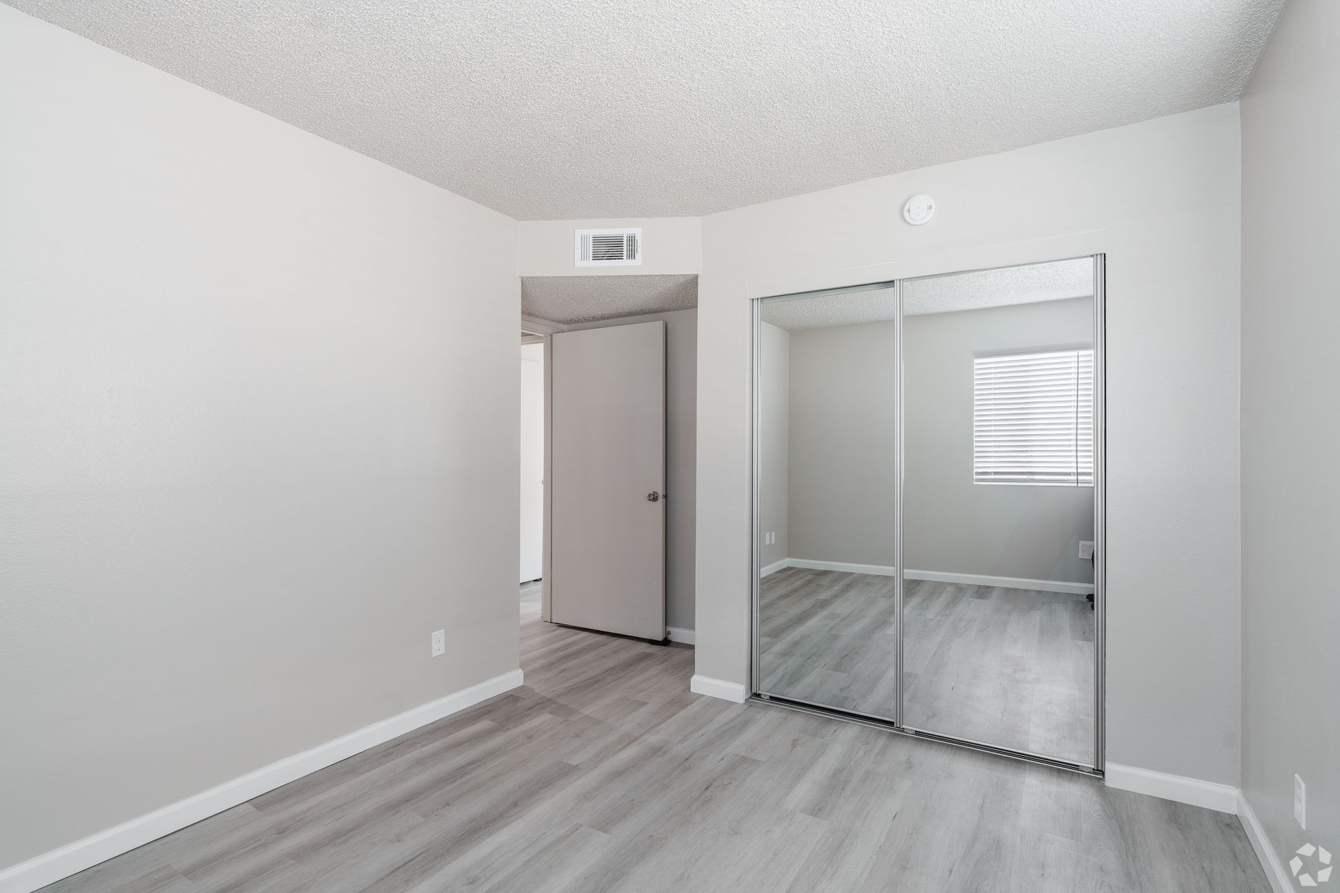 Empty bedroom with gray walls, mirrored closet doors, and wood-look flooring.