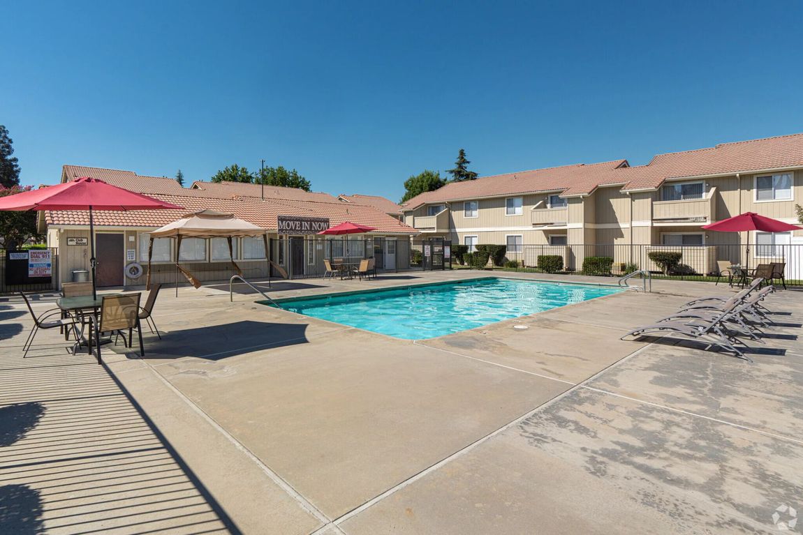 Swimming pool at apartment complex with lounge chairs, tables, and umbrellas under a blue sky.