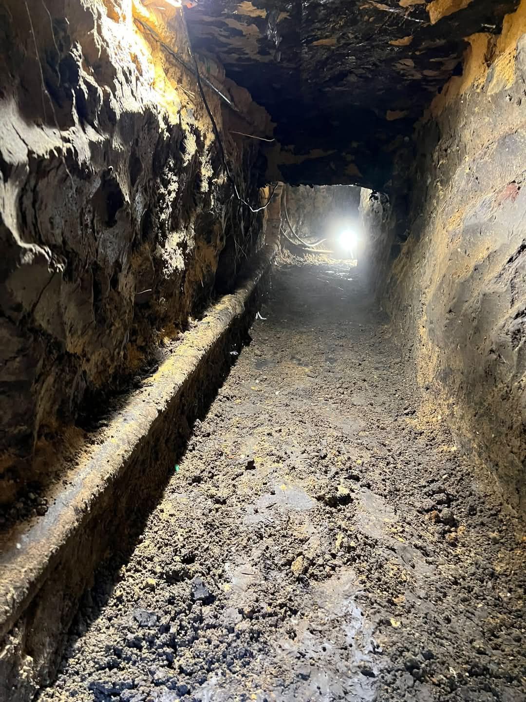 Tunnel with dirt walls and floor, light at the end, and a trench along the left.