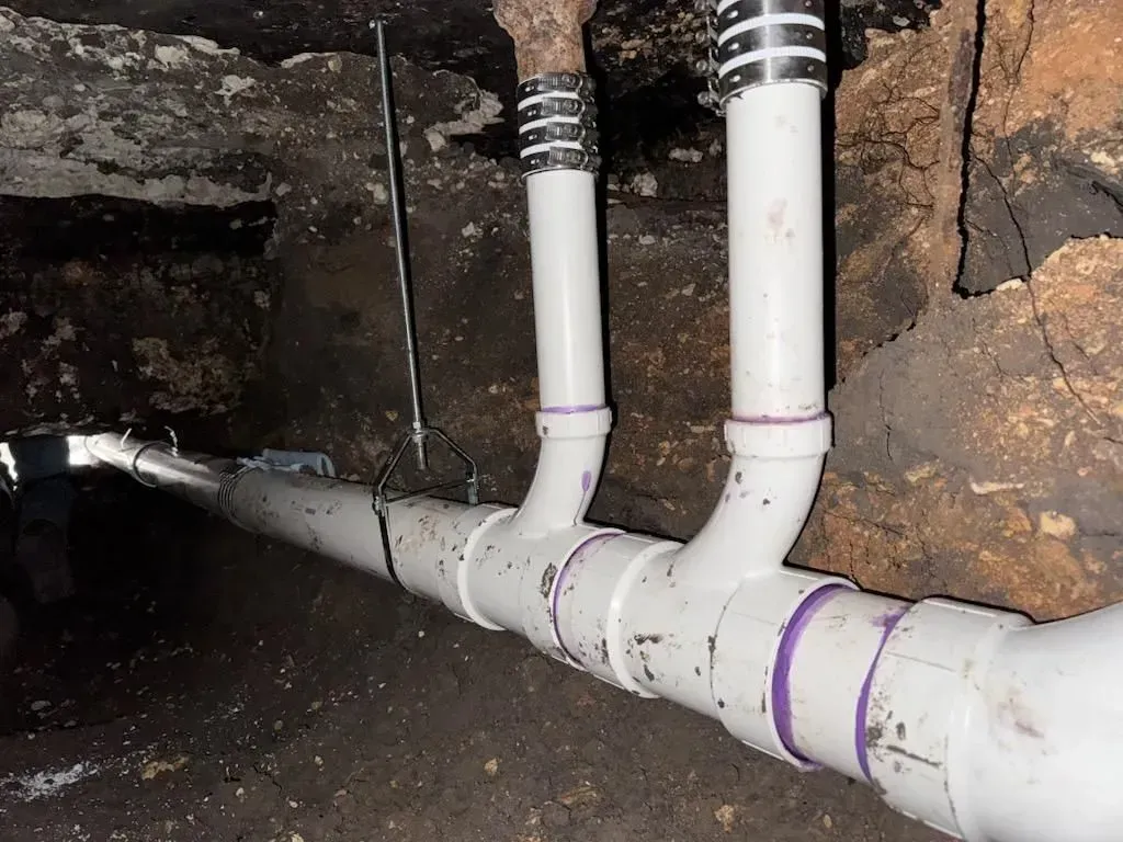 White PVC pipes with purple bands connect to vertical pipes in a dark crawl space.