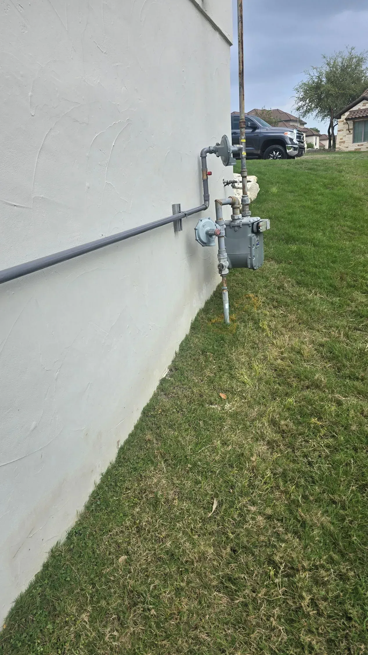 Gas meter and pipes on a white stucco wall next to a grassy lawn. A vehicle and trees are visible in the background.