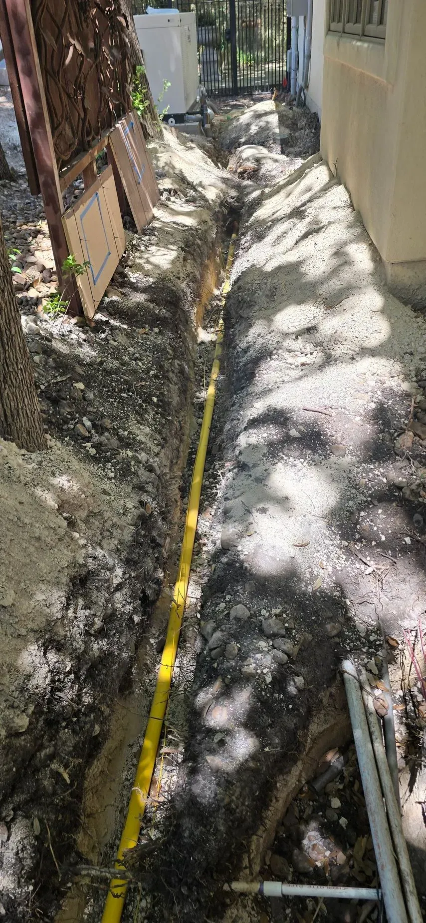 A narrow trench in the ground with a yellow pipe running along the bottom, along a wall.