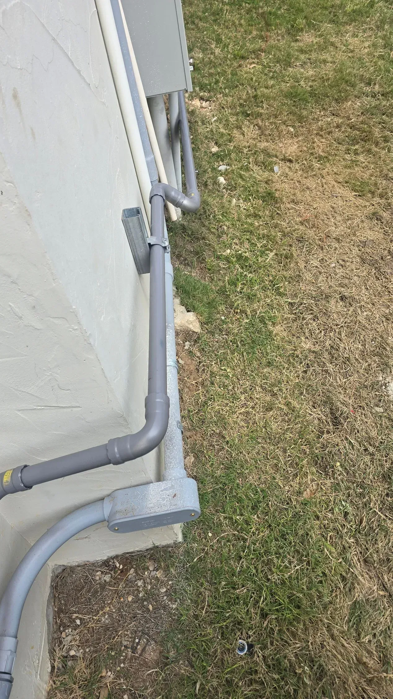Gray pipes attached to a light-colored wall, running down to a gray electrical box on the ground, beside grassy area.