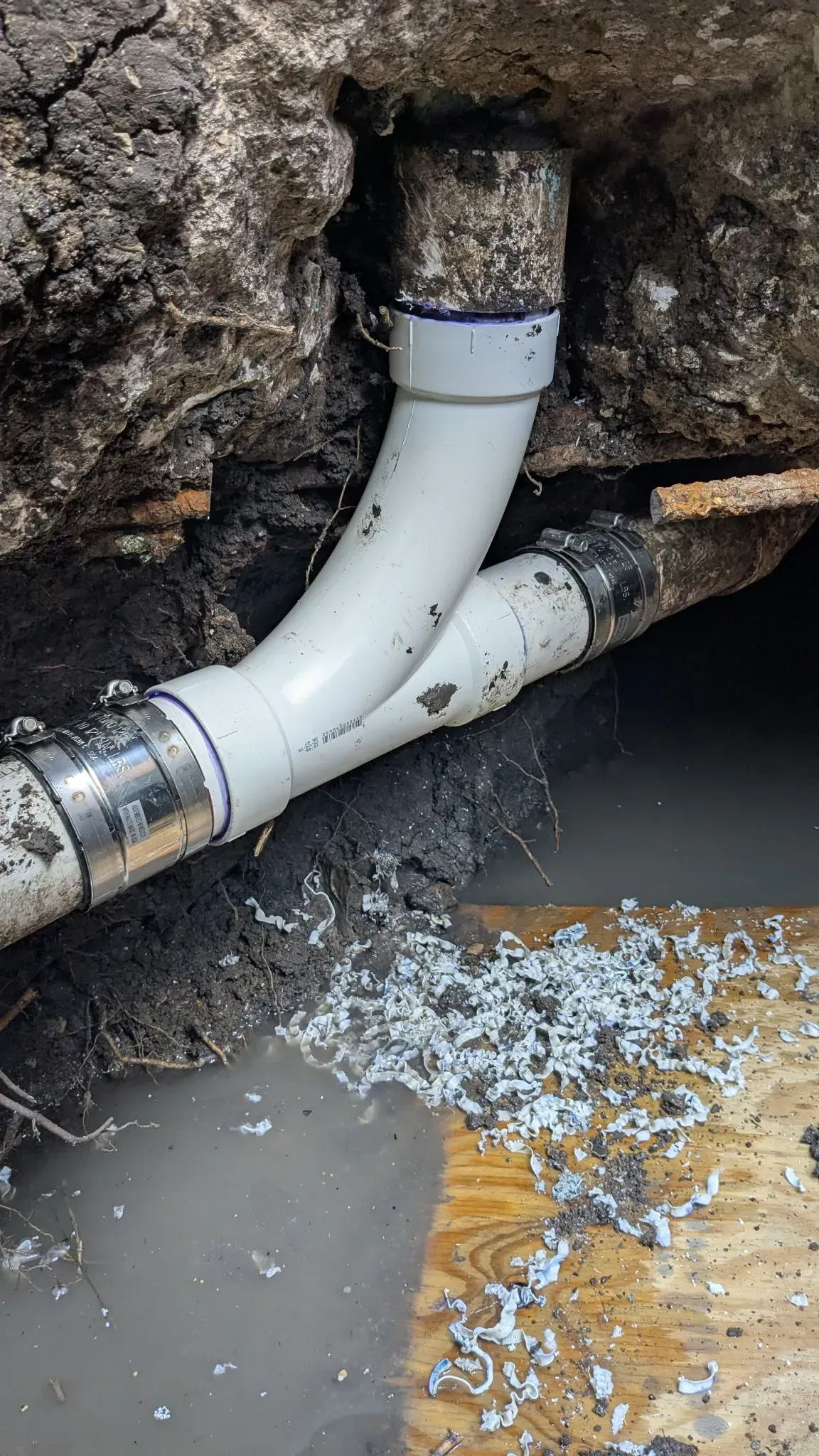 White PVC pipes and fittings in a dirt trench, connected to a metal pipe with clamps; some water visible.